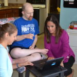 A man sits and watches as two staff members set up a laptop for a skype call
