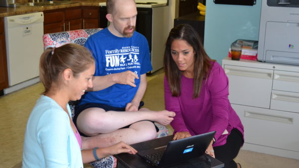 A man sits and watches as two staff members set up a laptop for a skype call