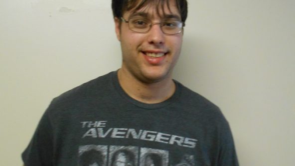 Ryan stands in front of a blank wall smiling. He is a young man wearing a grey Avengers shirt, and has brown shaggy hair and glasses.