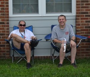 Two men sit in camping chairs at an outside event.