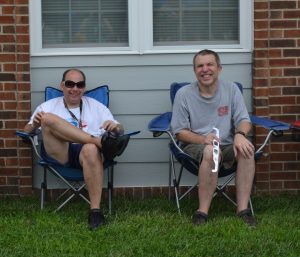 Two men sit in camping chairs at an outside event.