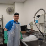 Bryan smiles at the camera while on duty at his dish washing job. He wears an apron and long rubber gloves while standing at a sink full of suds.