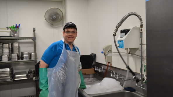Bryan smiles at the camera while on duty at his dish washing job. He wears an apron and long rubber gloves while standing at a sink full of suds.