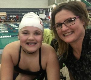 Sofia wears a swim cap and swimming suit and poses with her mom at a swim meet.