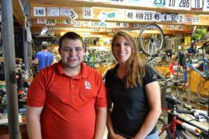 Aaron stands next to Sarah in the bike shop.