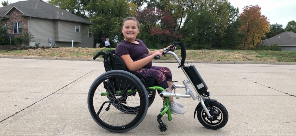 A teenage girl sits outdoors in an adapted wheelchair.