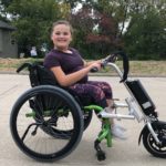A teenage girl sits outdoors in an adapted wheelchair.