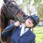 Woman wears horseback riding gear and smiles as she gets a nuzzle from her horse.