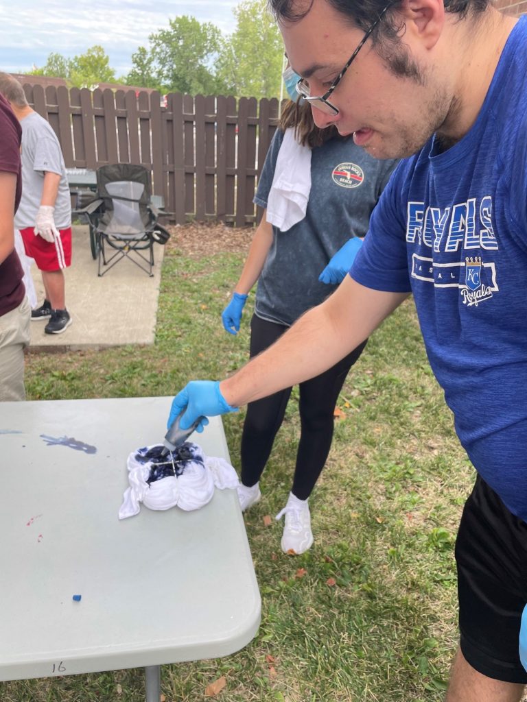 A man places blue dye onto a tied up t-shirt while standing outside at a table.