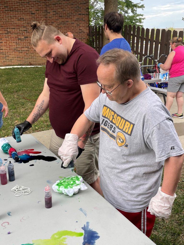 Two men place dye onto tied up t-shirts outdoors on a table.