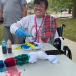 A woman smiles as she places various dyes onto a tied up t-shirt outdoors at a table.