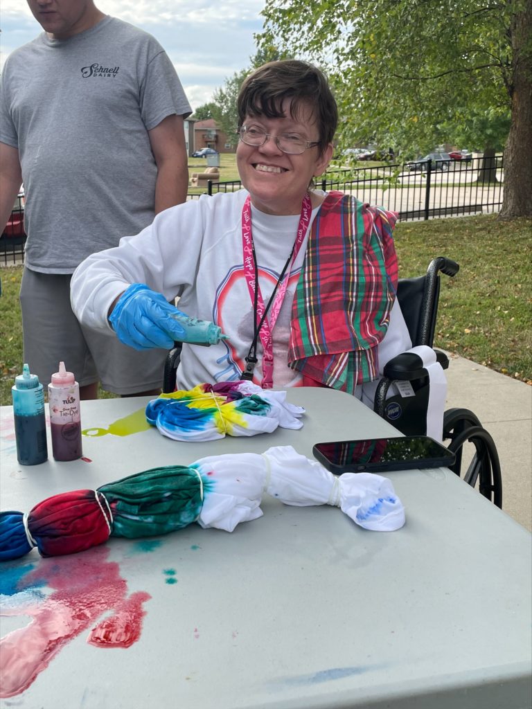A woman smiles as she places various dyes onto a tied up t-shirt outdoors at a table.