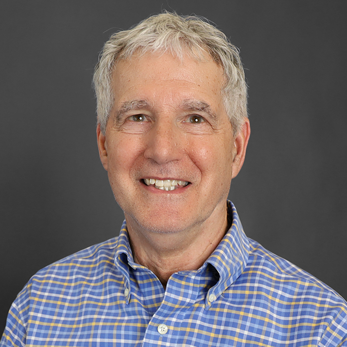 Bob is light skin man who is smiling brightly, wearing a light blue plaid oxford, light grey hair.