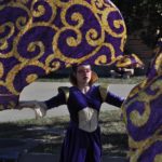 Girl dressed in purple costume waves a large ornate purple and gold flag.