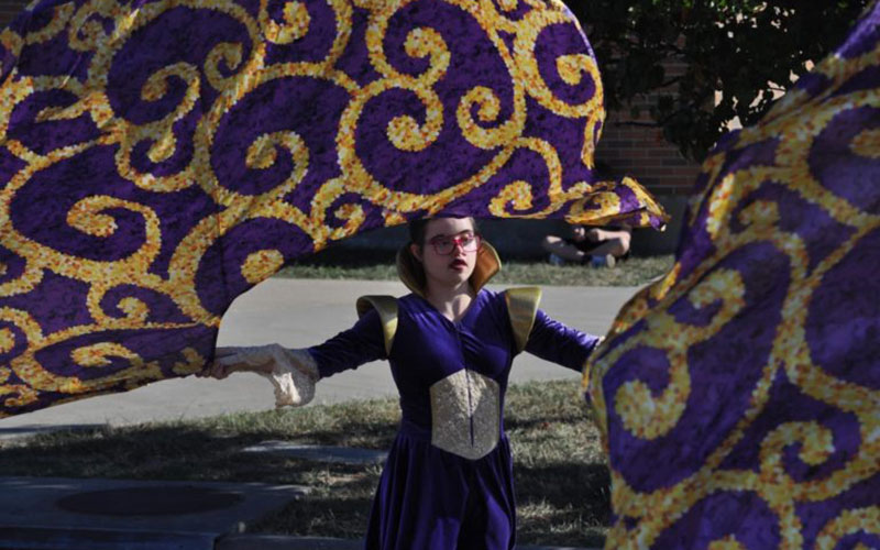 Girl dressed in purple costume waves a large ornate purple and gold flag.