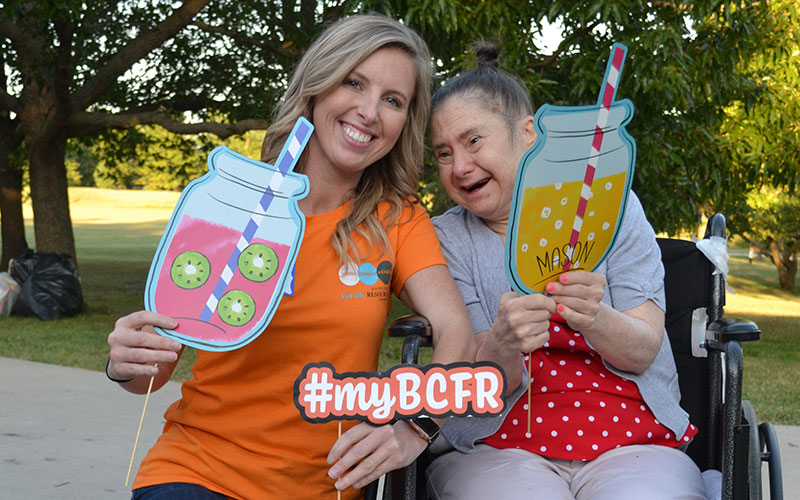 Hadyn wears a BCFR staff shirt in bright orange and kneels beside a woman in supported living who sits in a wheelchair. The women are smiling and holding fun summer cutouts in the shape of lemonade filled mason jars.