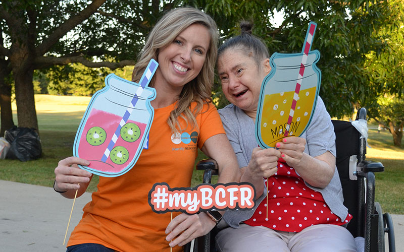 Hadyn wears a BCFR staff shirt in bright orange and kneels beside a woman in supported living who sits in a wheelchair. The women are smiling and holding fun summer cutouts in the shape of lemonade filled mason jars.