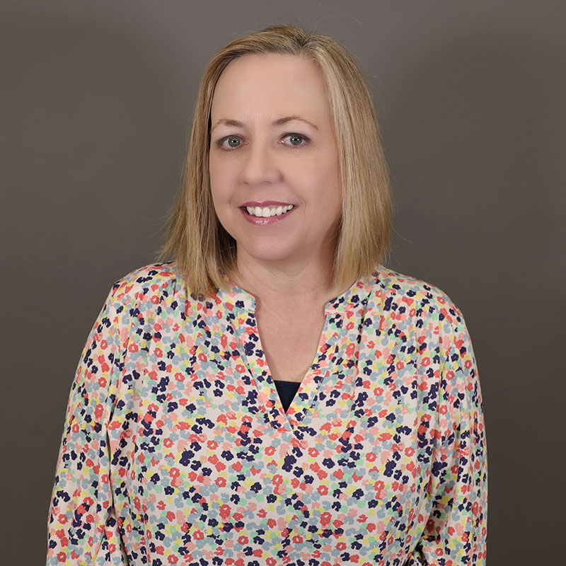 Laura Cravens is a light skin tone woman with blue eyes and medium-toned blonde hair cropped at her shoulders. She is smiling and wearing a brightly colored floral print shirt.