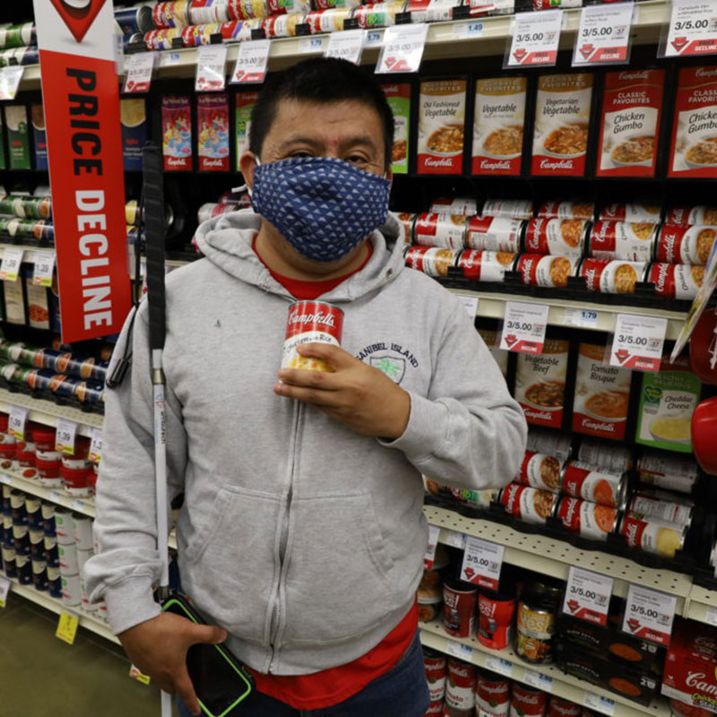 Tomas wears a face mask and holds a can of soup in one hand, and his phone and white cane in the other. Tomas has medium light skin tones, dark hair, and is wearing a zip up hoodie.