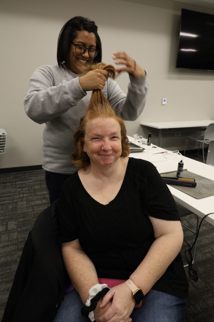 A woman with light skin and red hair is seated getting her hair curled.