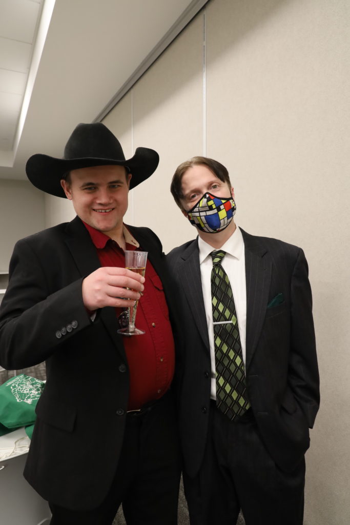 Two men dressed in suits pose for a photo. One man wears a cowboy hat and holds up a champagne class and the other man wears a suit, tye and colorful face mask.
