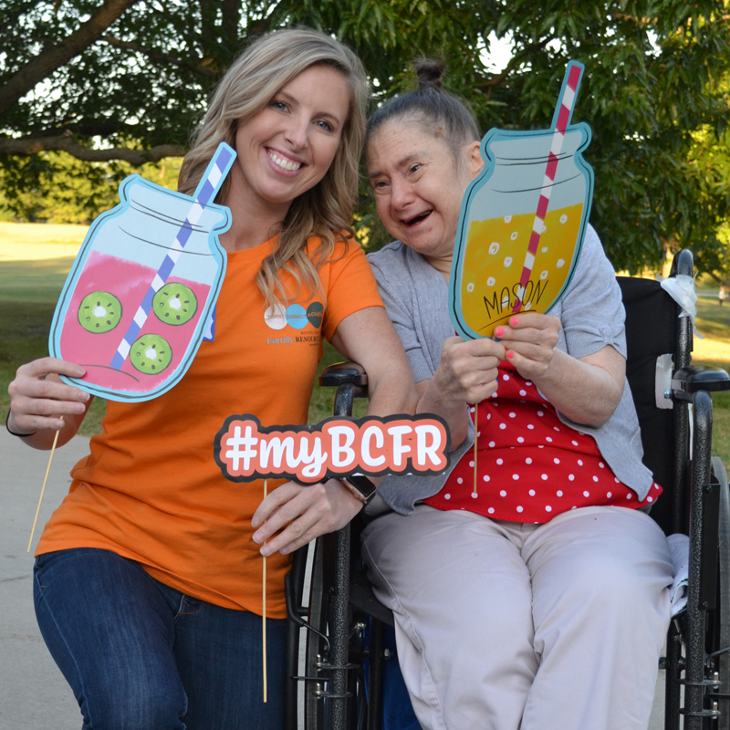 Hadyn wears a BCFR staff shirt in bright orange and kneels beside a woman in supported living who sits in a wheelchair. The women are smiling and holding fun summer cutouts in the shape of lemonade filled mason jars.