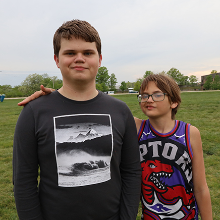 Two brothers stand on the track at West Elementary. Leo is on the left and is much taller than his younger brother Finn. Leo is a light skin male wearing a black shirt. Finn is a light skin male with a small frame, shaggy brown hair and glasses.