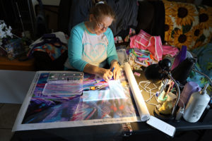 Tina sits at a table with a small lamp shining on her art canvas. She is placing diamonds into little squares. Tina is a light skin woman with red hair and glasses.