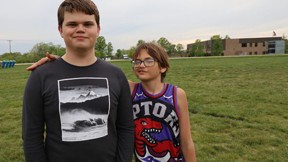 Two brothers stand on the track at West Elementary. Leo is on the left and is much taller than his younger brother Finn. Leo is a light skin male wearing a black shirt. Finn is a light skin male with a small frame, shaggy brown hair and glasses.