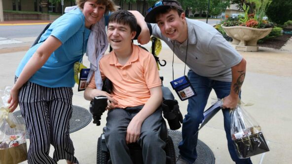 Three friends pose for a photo on Mizzou campus. A college age female and male stand on either side of Declan who is in a wheelchair wearing an orange polo and jeans.