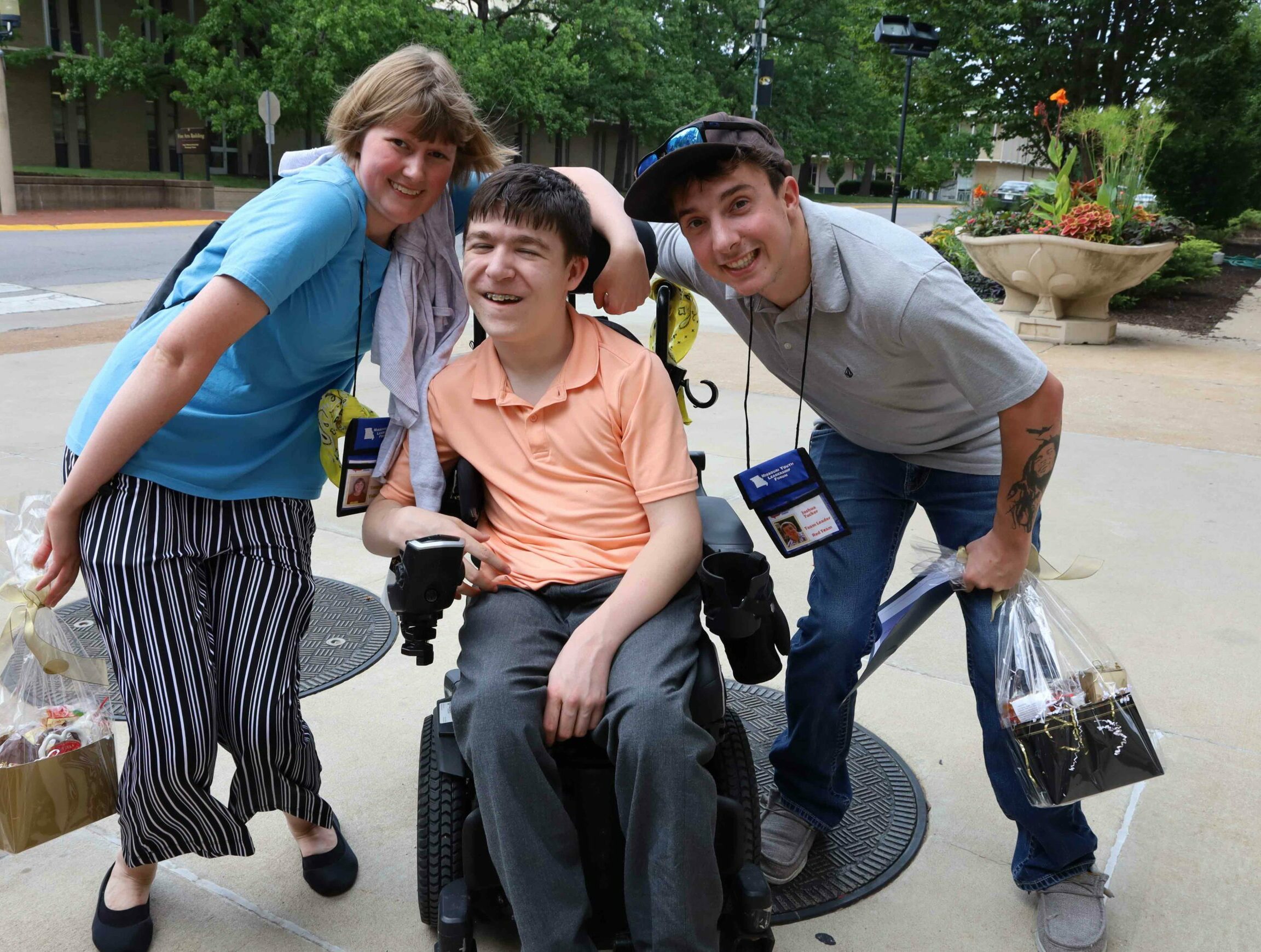 Three friends pose for a photo on Mizzou campus. A college age female and male stand on either side of Declan who is in a wheelchair wearing an orange polo and jeans.
