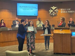 Lydia accepts an award at the October City Council meeting from Mayor Barbara Buffaloe. She stands near a podium holding a plaque and people stand and clap behind her.