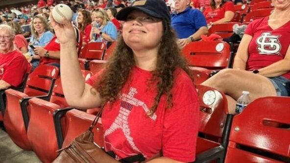 Lydia sits in a red seat at Busch Stadium holding a baseball proudly in the air. She wears a red shirt and blue baseball hat.