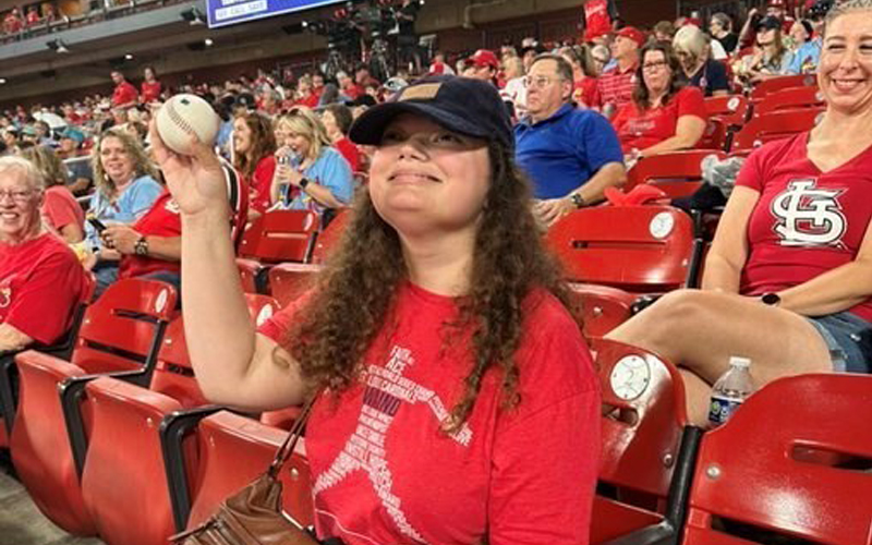 Lydia sits in a red seat at Busch Stadium holding a baseball proudly in the air. She wears a red shirt and blue baseball hat.