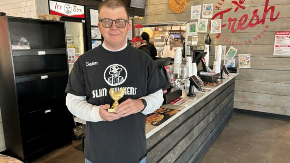 Galen holds a trophy shaped like a golden chicken in front of the cash register at Slim Chickens. He is wearing a black shirt with a Slim Chickens written in large white letters.