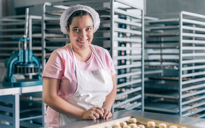 A woman in a bakery lays out cookie dough balls onto a sheetpan. She is wearing a pink shirt, white apron, and white hair net.