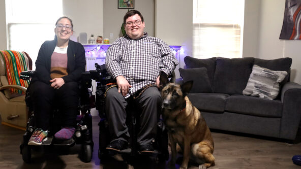 Lindsey, Colton and their dog Zara pose for a family photo in their living room in front of a fireplace and brown couch. Lindsey and Colton use wheelchairs.