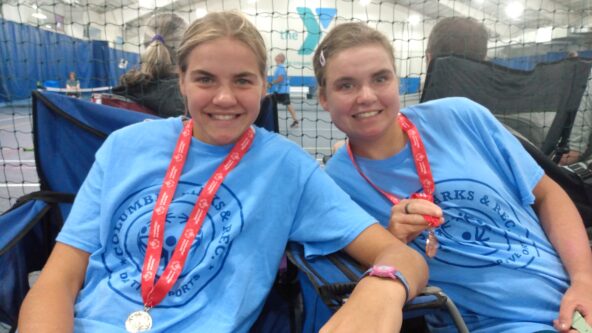 Two similar looking sisters wear blue special olympics shirts and medals around their necks.