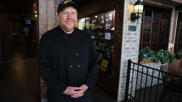 Tom stands on the indoor courtyard of Mort's in the MU Student Union. He is wearing a black baseball cap and black chef's coat.