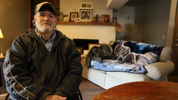 Joe sits at a table in his comfy new apartment. In the background is a tan coach and fireplace with framed photos on the mantle.
