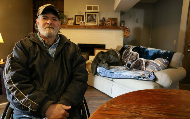 Joe sits at a table in his comfy new apartment. In the background is a tan coach and fireplace with framed photos on the mantle.