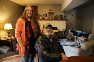 Joe sits a table in his quaint and cozy living room. His sister Angie stands behind him with her hand on his shoulder.