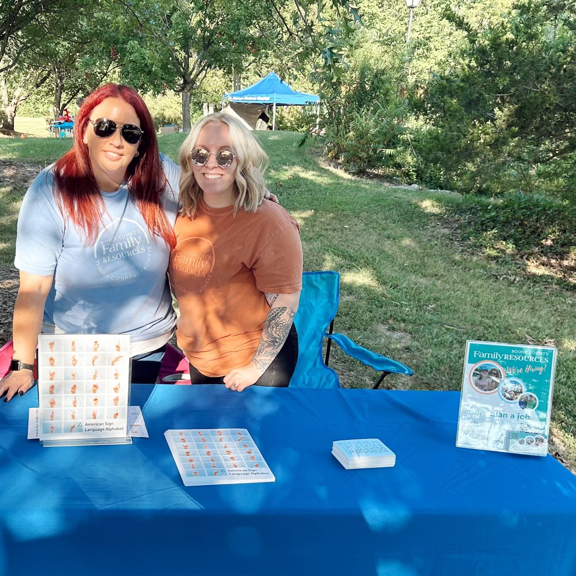 Two staff wearing BCFR T-shirts pose near a table filled with BCFR marketing items and an outdoor event.