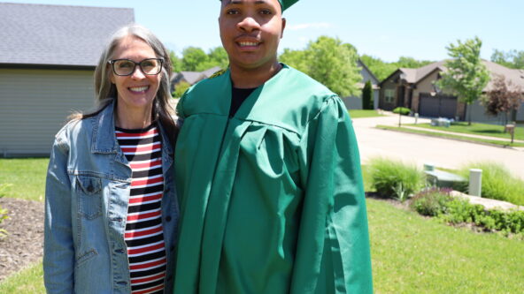 Drew wears a green cap and gown with a gold tassel. He is posing for a photo outside his house with his mom.