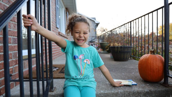Ayla has a big grin sitting on her porch next to a pumpkin. She is wearing bright clothes and has curly brown hair.
